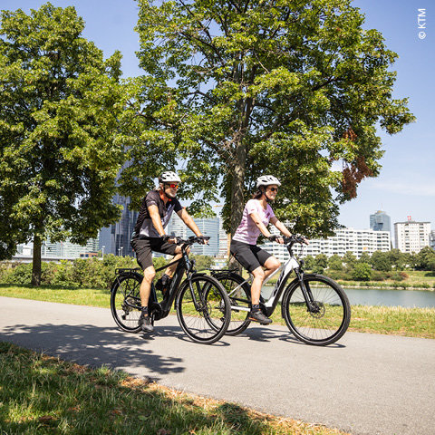 Zwei Personen fahren mit Helmen und legerer Radsportkleidung auf einem gepflasterten Weg durch einen Park. Im Hintergrund sind grüne Bäume und ein Gewässer zu sehen, in der Ferne sind Stadtgebäude unter einem klaren blauen Himmel zu erkennen.