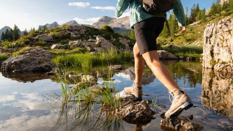 Eine Person in Wanderschuhen und mit Rucksack überquert einen steinigen Bach mit klarem Wasser. Die Szene spielt in einer Berglandschaft mit grünem Gras, verstreuten Felsen und Bäumen in der Ferne unter einem bewölkten Himmel.