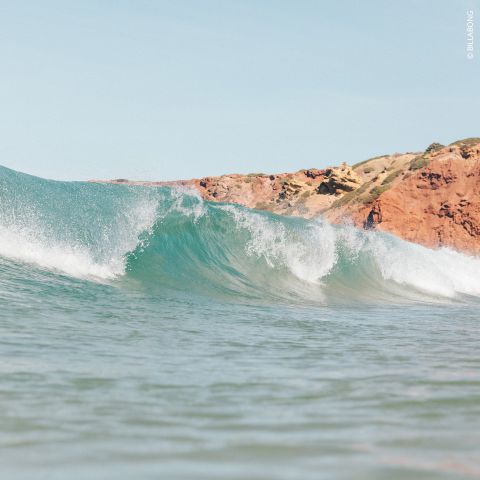 Meereswelle mit einer felsigen, rotbraunen Klippe im Hintergrund unter einem klaren blauen Himmel. Das Wasser ist türkis und schaumig. Sandstrand nicht sichtbar.