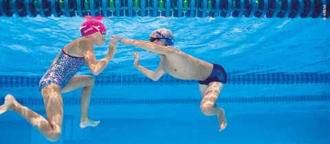 Zwei Kinder schwimmen unter Wasser in einem Pool. Sie stehen einander gegenüber, haben die Arme ausgestreckt und tragen Badekappen und Schwimmbrillen. Das Wasser ist klar und über ihnen sind die Bahnbegrenzungen des Pools zu sehen.