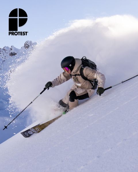 Ein Skifahrer in beiger Ausrüstung und Helm fährt durch den Neuschnee an einem steilen Hang und hinterlässt eine Schneewolke. Das Logo „PROTEST“ ist in der oberen linken Ecke zu sehen. Im Hintergrund sind ein klarer Himmel und schneebedeckte Berge zu sehen.