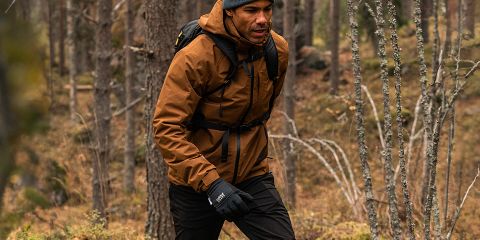 Mann in brauner Jacke, schwarzer Hose und grauer Mütze wandert durch einen dichten Wald. Er trägt schwarze Handschuhe und einen Rucksack. Der Wald hat hohe Bäume und abgefallene Blätter. „Rukka“-Logo oben.
