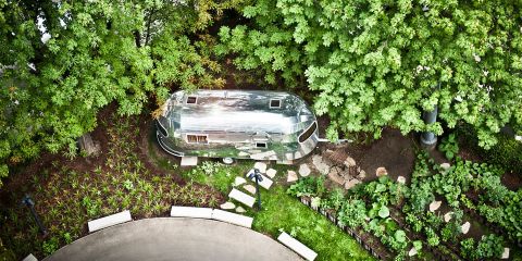 Ein silberner Airstream-Wohnwagen steht in einem üppigen Garten, der von dichten grünen Bäumen umgeben ist. Der Bereich umfasst einen kleinen Pfad mit Trittsteinen und weißen, geschwungenen Bänken im Vordergrund, von oben betrachtet.