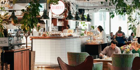 Ein gemütliches Café-Interieur mit Leuten, die an Tischen sitzen. Die Bäckereitheke ist mit Gebäck gefüllt und ein Barista bereitet Getränke zu. Der Raum ist mit Grünpflanzen und stimmungsvollem Licht dekoriert, was eine warme und einladende Atmosphäre schafft.
