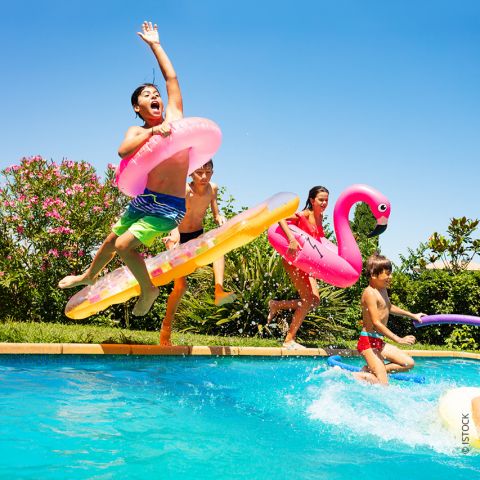 Kinder springen freudig in einen Swimmingpool und halten dabei aufblasbare Spielzeuge in der Hand, darunter einen rosa Flamingo und einen aufblasbaren Donut. Der klare blaue Himmel und das üppige Grün sorgen für eine lebendige, fröhliche Atmosphäre.