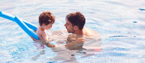 Ein Mann steht in einem Schwimmbecken und hält ein Kind im Arm. Das Kind hält eine blaue Schwimmnudel. Das Wasser ist klar und beide scheinen ihre Zeit im Becken zu genießen.