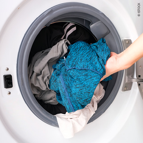 Eine Hand legt ein blaues Kleidungsstück in eine Waschmaschine. Die Maschine ist bereits mit anderen Kleidungsstücken in verschiedenen Farben gefüllt, darunter Schwarz und Grau. Die Szene wird von oben betrachtet, mit Fokus auf die offene Tür des Geräts.