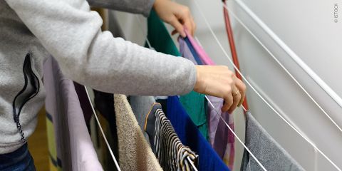 Person hängt Wäsche auf einen Wäscheständer. Zu sehen sind verschiedenfarbige Kleidungsstücke, darunter gestreifte und einfarbige Muster. Die Person trägt eine graue Jacke und ist von der Seite zu sehen, konzentriert auf das Ordnen der Kleidung.