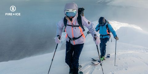 Zwei Personen in Winterkleidung fahren auf Skiern einen Berg hinauf durch den Schnee. Sie tragen Rucksäcke und nutzen Stöcke zur Unterstützung. Im Hintergrund ist eine neblige, weite Landschaft zu sehen. Das Bild enthält in der oberen linken Ecke ein „Fire + Ice“-Logo.