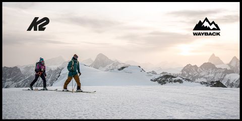 Zwei Personen fahren unter einem bewölkten Himmel einen schneebedeckten Berg hinauf, im Hintergrund schroffe Gipfel. Die Logos von K2 und Wayback befinden sich in den oberen Ecken.