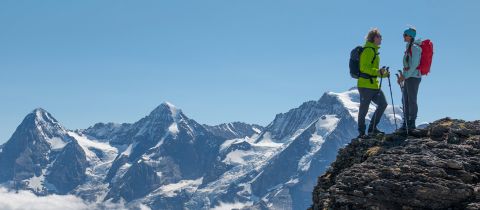 Zwei Wanderer in hellen Jacken und Rucksäcken stehen auf einem felsigen Bergkamm und blicken auf eine Reihe schneebedeckter Gipfel unter einem klaren blauen Himmel.