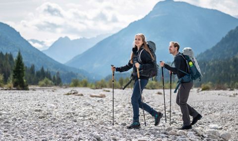 Eine Frau und ein Mann wandern mit Rucksäcken und Trekkingstöcken durch felsiges Gelände. Sie tragen Outdoor-Ausrüstung. Im Hintergrund sind Berge und ein bewölkter Himmel zu sehen. Die Szene ist hell und malerisch.