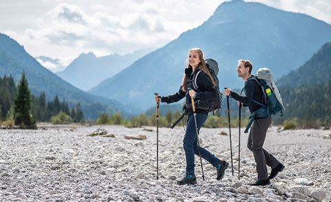 Eine Frau und ein Mann wandern mit Rucksäcken und Trekkingstöcken durch felsiges Gelände. Sie tragen Outdoor-Ausrüstung. Im Hintergrund sind Berge und ein bewölkter Himmel zu sehen. Die Szene ist hell und malerisch.