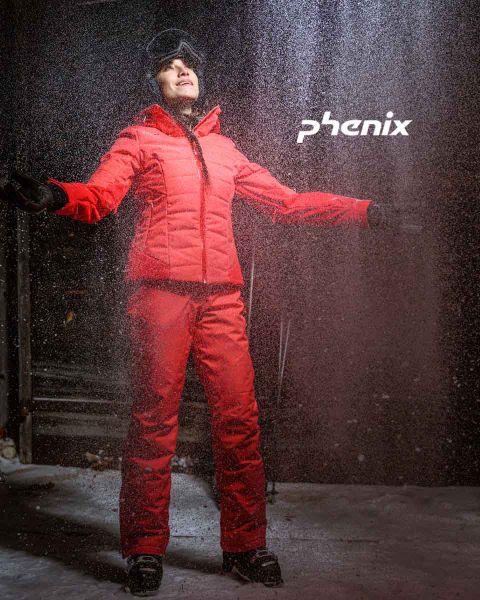 Eine Person steht draußen im Schnee. Sie trägt ein leuchtend rotes Ski-Outfit und schwarze Handschuhe. Sie hat die Arme ausgestreckt und blickt nach oben. Über ihr ist das Wort „Phenix“ zu sehen. Die Szene lässt darauf schließen, dass die Person das Winterwetter genießt.