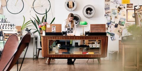 Eine Person steht hinter einer Holztheke in einem Geschäft im Vintage-Stil. An den Wänden hängen Fahrräder, Pflanzen, Fotos und Schallplatten. Die Person ist konzentriert und um die Theke herum stehen verschiedene Gegenstände wie Tassen und Flaschen.
