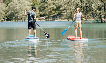 Zwei Personen paddeln auf einem ruhigen See. Der Mann links steht auf einem blauen Board, während die Frau rechts auf einem roten Board steht. Sie sind an einem sonnigen Tag von Bäumen und klarem Wasser umgeben.