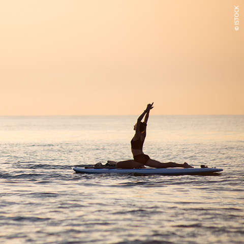 Eine Person praktiziert Yoga auf einem Paddleboard im ruhigen Ozean. Sie führt eine sitzende Vorwärtsbeuge mit erhobenen Armen aus, die Silhouette steht vor einem warmen Sonnenuntergangshimmel.