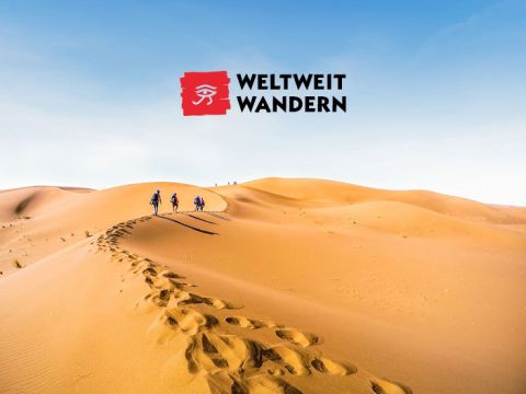 Menschen wandern unter einem klaren blauen Himmel auf Sanddünen. Oben links steht ein Logo mit der Aufschrift „Weltweit Wandern“ und einem stilisierten Augensymbol. Hinter den Wanderern sind Fußspuren zu sehen, die die weite, offene Wüstenlandschaft betonen.