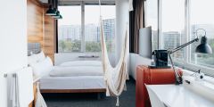 Modernes Hotelzimmer mit großen Fenstern und Blick auf die Stadt. Das Zimmer verfügt über ein Doppelbett, weiße Bettwäsche, eine Hängematte, eine Schreibtischlampe, eine Kaffeemaschine und einen Fernseher. Die Einrichtung umfasst Holzakzente und einen Arbeitsbereich mit einem weißen Schreibtisch.