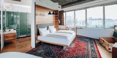 Ein modernes Hotelzimmer mit einem großen Bett auf einem rot gemusterten Teppich. Das Zimmer verfügt über Holzakzente, Hängelampen, eine Dusche mit Glaswänden und große Fenster mit Blick auf die Stadt. Ein kleiner Schreibtisch und ein Stuhl stehen neben dem Fenster.