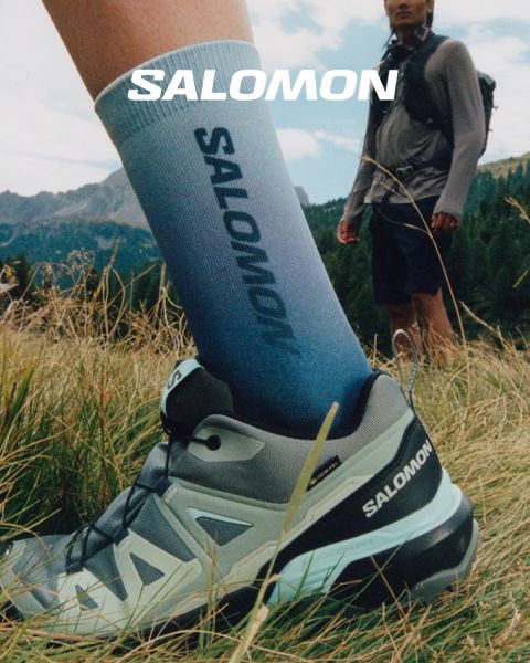 Nahaufnahme einer Person in Salomon-Schuhen und Salomon-Socken mit Farbverlauf in Blau. Sie steht auf einer Wiese mit einer malerischen Berglandschaft im Hintergrund. Ein Wanderer in Outdoor-Kleidung steht weiter entfernt, ebenfalls auf der Wiese.