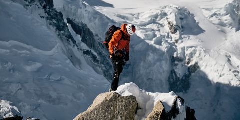 Ein Kletterer in orangefarbener Jacke und Helm steht auf schneebedeckten Felsen vor einer Kulisse schroffer, eisiger Berge. Er ist mit Kletterausrüstung, darunter Klettergurt und Seilen, ausgestattet und blickt auf seine Füße.