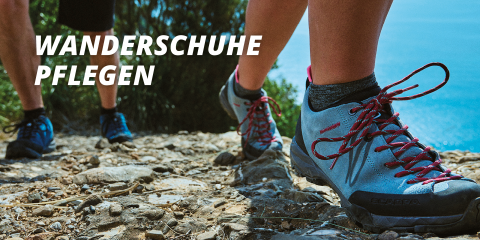 Nahaufnahme von zwei Personen, die auf einem steinigen Pfad wandern. Der Fokus liegt auf ihren Schuhen. Die Person im Vordergrund trägt graue Wanderschuhe mit roten Schnürsenkeln. Der Text auf dem Bild lautet „WANDERSCHUHE PFLEGEN“. Im Hintergrund sind blaues Wasser und grünes Laub zu sehen.