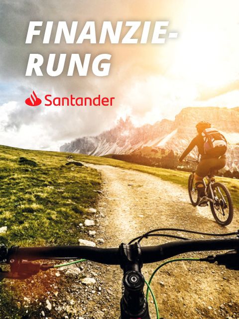 Zwei Personen radeln auf einem Schotterweg durch eine Berglandschaft mit dramatischen Wolken und Sonnenlicht. In der oberen linken Ecke sind das Wort „Finanzierung“ und das Santander-Logo zu sehen.