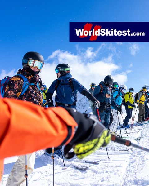 Eine Gruppe Skifahrer in farbenfroher Ausrüstung steht mit Skistöcken, Helmen und Schutzbrillen auf einem verschneiten Berghang. Der Himmel ist klar und blau. Oben rechts steht ein großes Logo mit der Aufschrift „WorldSkitest.com“.