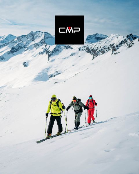 Drei Skifahrer in farbenfrohen Outfits erklimmen unter einem klaren blauen Himmel einen schneebedeckten Berg, umgeben von hohen, schneebedeckten Gipfeln. Das schwarze Logo „CMP“ erscheint oben in der Mitte des Bildes.