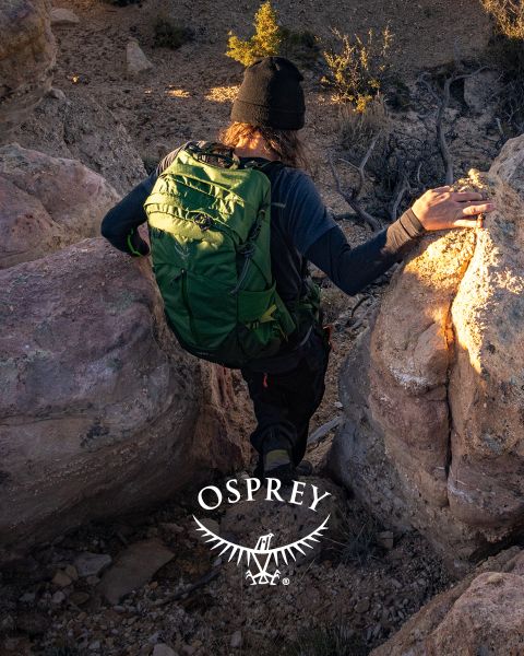 Eine Person steigt mit einem grünen Rucksack, einer schwarzen Mütze und dunkler Kleidung ein felsiges Gelände hinab. Sonnenlicht beleuchtet die Landschaft. Im Vordergrund ist das Osprey-Logo zu sehen.