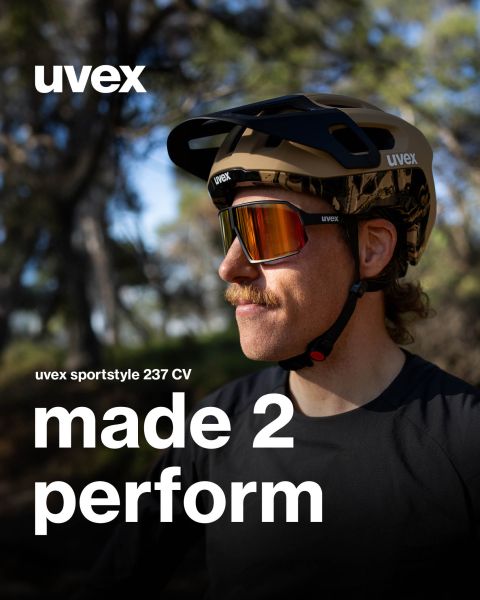 Eine Person mit Schnurrbart trägt eine UVEX Sportstyle-Brille und einen Helm im Freien. Der Text lautet: „UVEX sportstyle 237 CV made 2 perform“. Im Hintergrund sind Bäume und verschwommenes Grün zu sehen.
