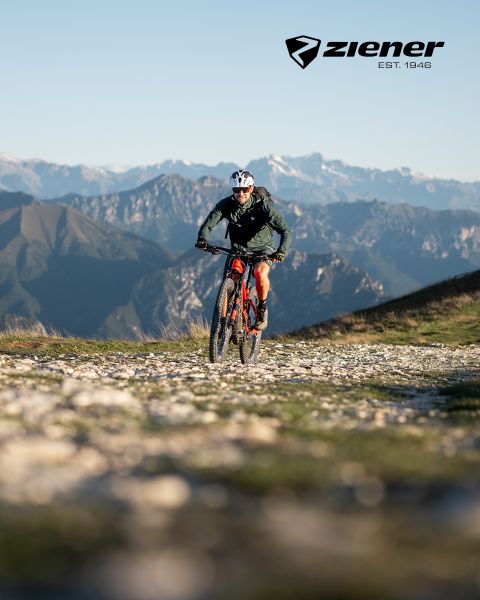 Ein Radfahrer fährt mit seinem roten Mountainbike auf einem Schotterweg vor der Kulisse schroffer Berge. Der Himmel ist klar, der Radfahrer trägt Helm und grüne Jacke. Oben rechts befindet sich das Ziener-Logo.