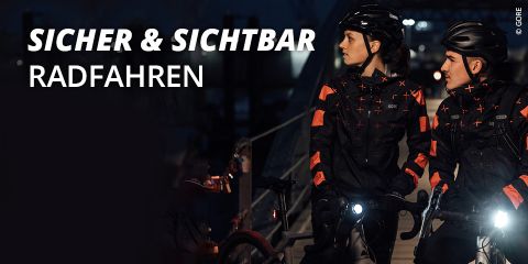 Zwei Radfahrer in reflektierender Ausrüstung sitzen nachts auf Fahrrädern. Sie tragen Helme und Jacken mit reflektierenden Streifen, während Fahrradlichter die Szene erhellen. Der große Text links lautet „Sicher & Sichtbar Radfahren“.
