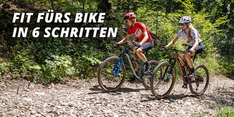 Zwei Radfahrer fahren mit Mountainbikes auf einem steinigen Pfad durch üppiges Grün. Beide tragen Helme und Sonnenbrillen. Der Text auf dem Bild lautet „FIT FÜRS BIKE IN 6 SCHRITTEN“. Die Szene ist sonnig und unterstreicht ein Outdoor-Radabenteuer.