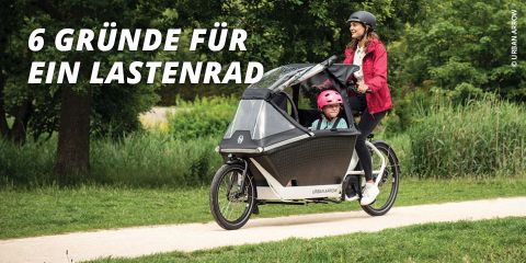 Eine Frau mit Helm fährt mit einem Lastenrad auf einem Weg durch einen Park. Ein Kind mit einem rosa Helm sitzt vorne im Fahrradfach. Der Text lautet „6 Gründe für ein Lastenrad“ mit einem Baum und Gras im Hintergrund.