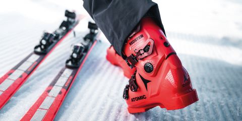 Eine Person mit leuchtend roten Skischuhen der Marke „Atomic“ steht neben roten und schwarzen Skiern auf einer schneebedeckten Oberfläche. Die Nahaufnahme fängt die Details der Schnallen der Schuhe und den strukturierten Schnee darunter ein.