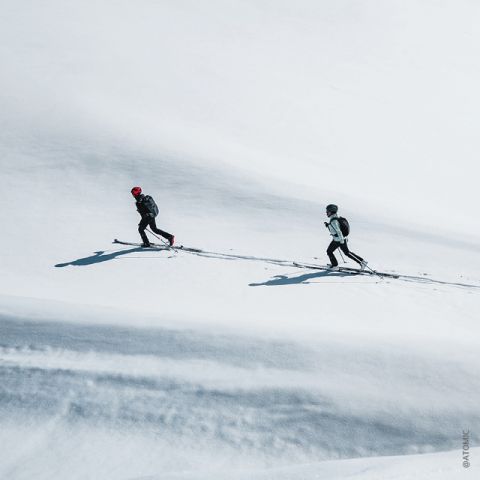 Zwei Skifahrer in schwarzer Skiausrüstung und Helmen fahren in einer verschneiten Landschaft bergauf. Der Skifahrer vorn trägt eine rote Mütze. Im Hintergrund ist eine weite Fläche unberührten weißen Schnees zu sehen.