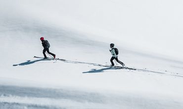 Zwei Skifahrer in Winterkleidung fahren einen schneebedeckten Hang hinauf. Der Skifahrer vorn trägt einen roten Helm, der hinter ihnen einen schwarzen. Beide werfen lange Schatten auf den weißen Schnee, während sie bergauf fahren.