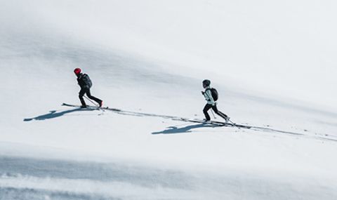 Zwei Skifahrer in Winterkleidung fahren einen schneebedeckten Hang hinauf. Der Skifahrer vorn trägt einen roten Helm, der hinter ihnen einen schwarzen. Beide werfen lange Schatten auf den weißen Schnee, während sie bergauf fahren.