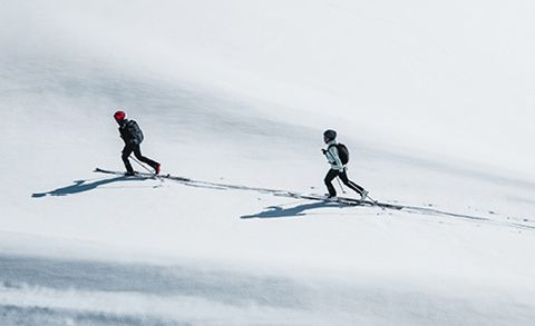 Zwei Skifahrer in Winterkleidung fahren einen schneebedeckten Hang hinauf. Der Skifahrer vorn trägt einen roten Helm, der hinter ihnen einen schwarzen. Beide werfen lange Schatten auf den weißen Schnee, während sie bergauf fahren.