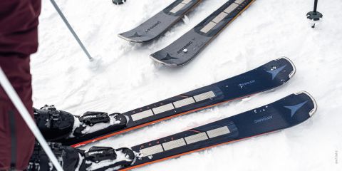 Nahaufnahme einer Person in Skistiefeln, die auf schwarzen Atomic-Skiern im Schnee steht. Zwei Paar Skier liegen daneben, und auch Skistöcke sind vorhanden. Die Szene fängt eine verschneite Winterlandschaft ein.
