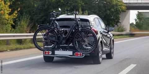 Ein silbernes Auto fährt auf einer Autobahn mit zwei Fahrrädern, die auf einem hinteren Fahrradträger montiert sind. Die Straße ist von Bäumen gesäumt und im Hintergrund ist eine große Betonbrücke zu sehen. Die Szene suggeriert eine Reise oder einen Ausflug.