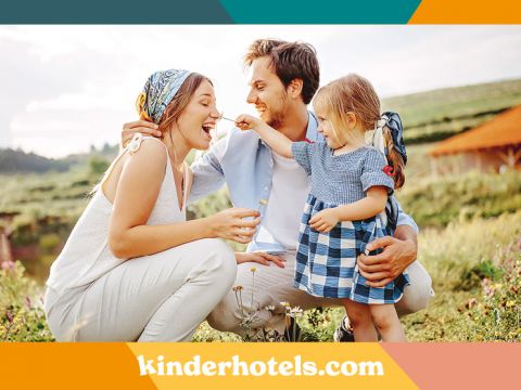 Eine dreiköpfige Familie genießt die Zeit im Freien auf einer Wiese. Frau und Mann hocken neben ihrer kleinen Tochter, die verspielt eine Blume hält. Alle lächeln und sind leger gekleidet. Unten ist der Text „kinderhotels.com“ zu sehen.