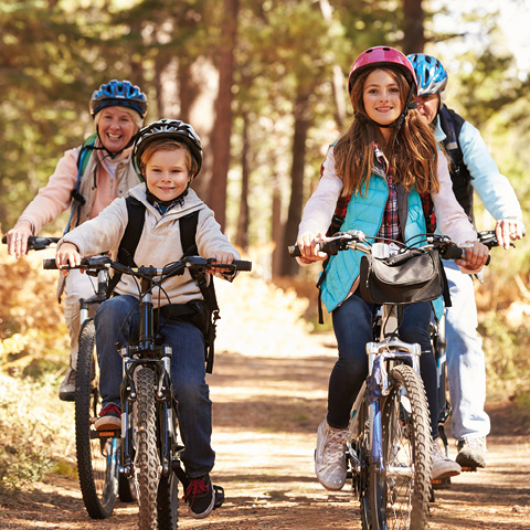 Eine vierköpfige Familie radelt auf einem Waldweg. Ein junges Mädchen und ein Junge gehen voran, beide mit Helmen. Hinter ihnen sind zwei Erwachsene, ebenfalls mit Helmen, die lächeln, während sie durch den sonnenbeschienenen Wald fahren. Alle scheinen die Outdoor-Aktivität zu genießen.