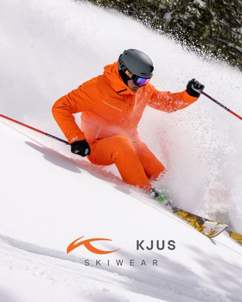 Ein Skifahrer in leuchtend orangefarbener Skiausrüstung und schwarzem Helm fährt durch den frischen Schnee und wirbelt dabei Pulverschnee auf. Der Hintergrund ist verschneit, und im unteren Teil des Bildes ist das Logo von KJUS Skiwear zu sehen.