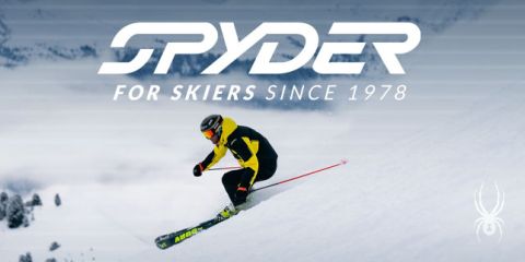 Ein Skifahrer in schwarz-gelbem Outfit fährt einen schneebedeckten Hang hinunter, im Hintergrund eine schneebedeckte Landschaft. Oben steht der Text: „SPYDER FÜR SKIFAHRER SEIT 1978.“ In der unteren rechten Ecke ist ein kleines weißes Spinnenlogo zu sehen.