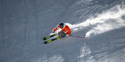 Ein Skifahrer in gelb-schwarzem Outfit und Helm legt sich auf einem schneebedeckten Hang scharf in eine Kurve und spritzt dabei Schnee auf. Der Himmel ist klar und die Ausrüstung des Skifahrers ist mit dem Wort „Fischer“ gekennzeichnet.