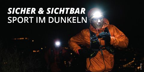 Zwei Personen laufen nachts mit Stirnlampen und orangefarbenen Jacken. Der Text lautet „SICHER & SICHTBAR SPORT IM DUNKELN“. Der Hintergrund ist eine verschwommene Stadtlandschaft mit Lichtern.