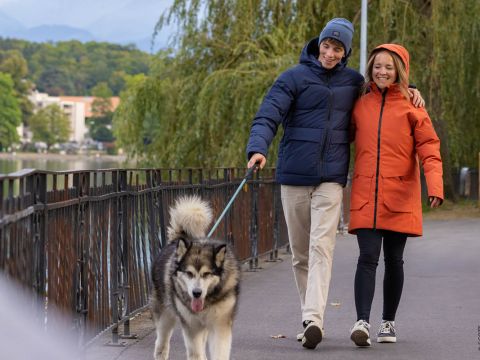 Ein Paar in Jacken führt seinen flauschigen Hund an der Leine einen von Bäumen gesäumten Uferweg entlang. Der Mann in blauer Jacke und Mütze hat seinen Arm um die Frau im orangefarbenen Mantel gelegt. Der Hund führt fröhlich den Weg.